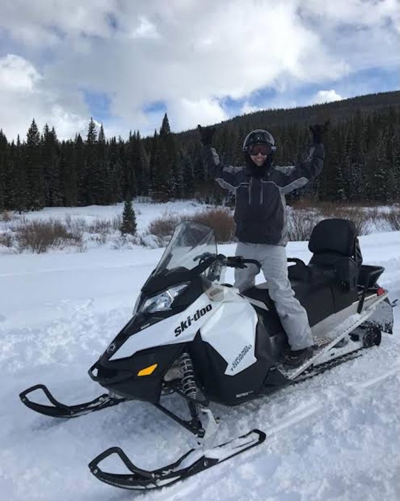 Eine Person steht auf einem Schneemobil, trägt Helm und Jacke, in einer verschneiten Landschaft mit Bäumen im Hintergrund.