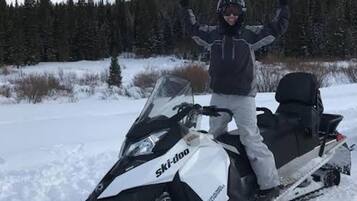 Eine Person steht auf einem Schneemobil, trägt Helm und Jacke, in einer verschneiten Landschaft mit Bäumen im Hintergrund.