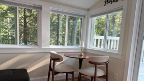 Una habitación con una mesa de comedor y sillas, ventanas amplias con vistas a árboles y un sofá de color oscuro.