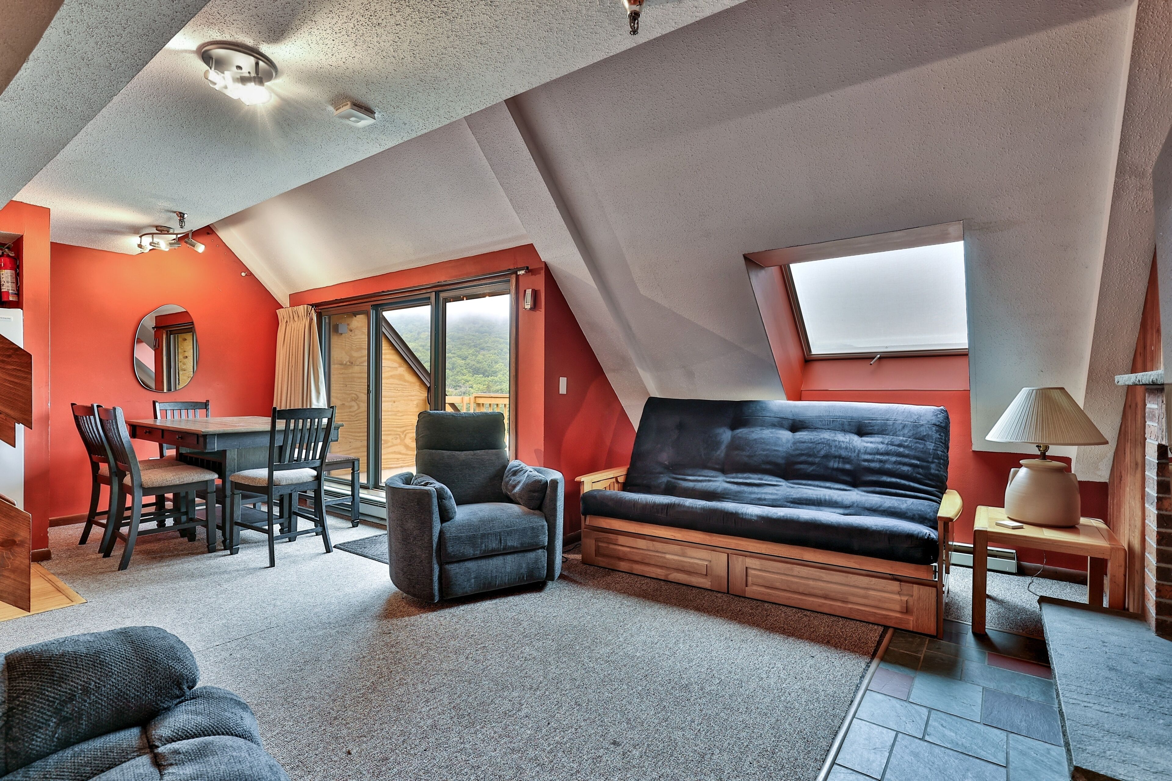 A cozy loft bedroom with a sloped ceiling, a sofa bed, a dining table with chairs, and a large window offering a view.