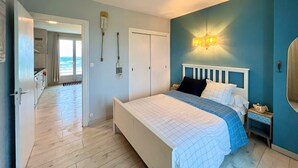 Quarto com cama, mesa de cabeceira, luminária na parede, porta e vista do oceano pela janela.