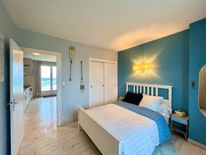 Een slaapkamer met een bed, een nachtkastje, een wandlamp, een deur en uitzicht op de oceaan door een raam.