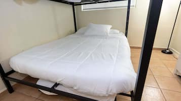 A single bed with a metal frame, white bedding, and a window with blinds in the background.