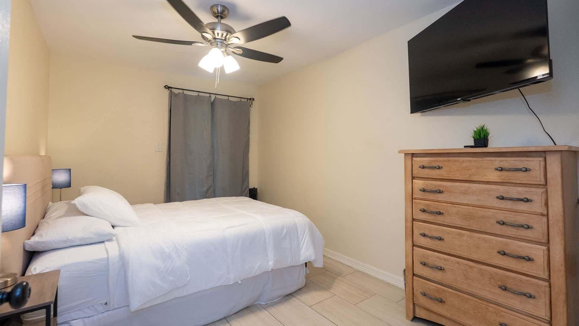 Een slaapkamer met een bed, een plafondventilator, een televisie en een houten dressoir.