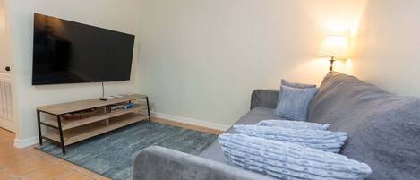 A living room with a flat-screen TV, a wooden TV stand, a grey sofa, and a lamp.