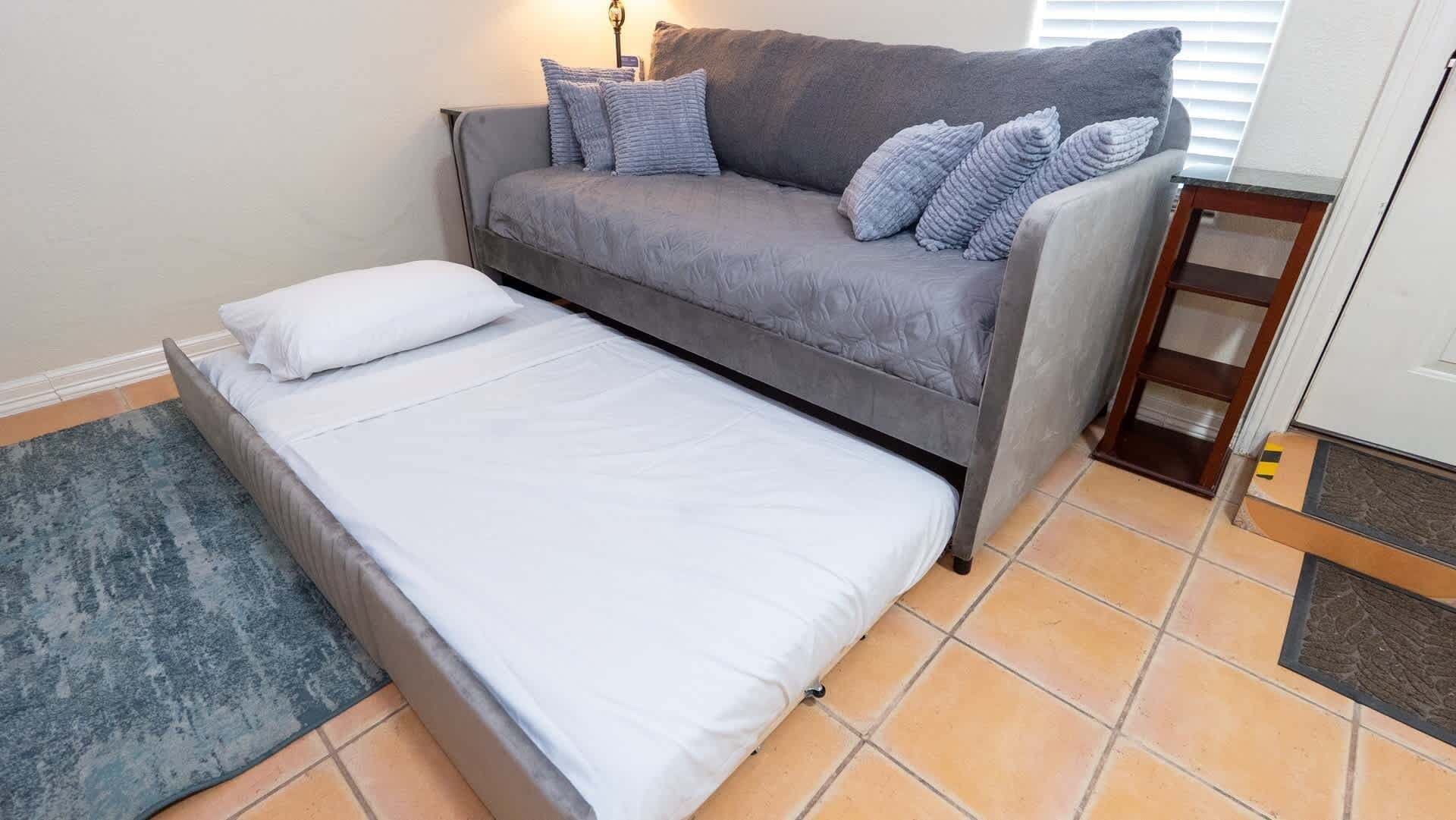 A compact room with a grey sofa, a single bed, a small side table, and a tiled floor.