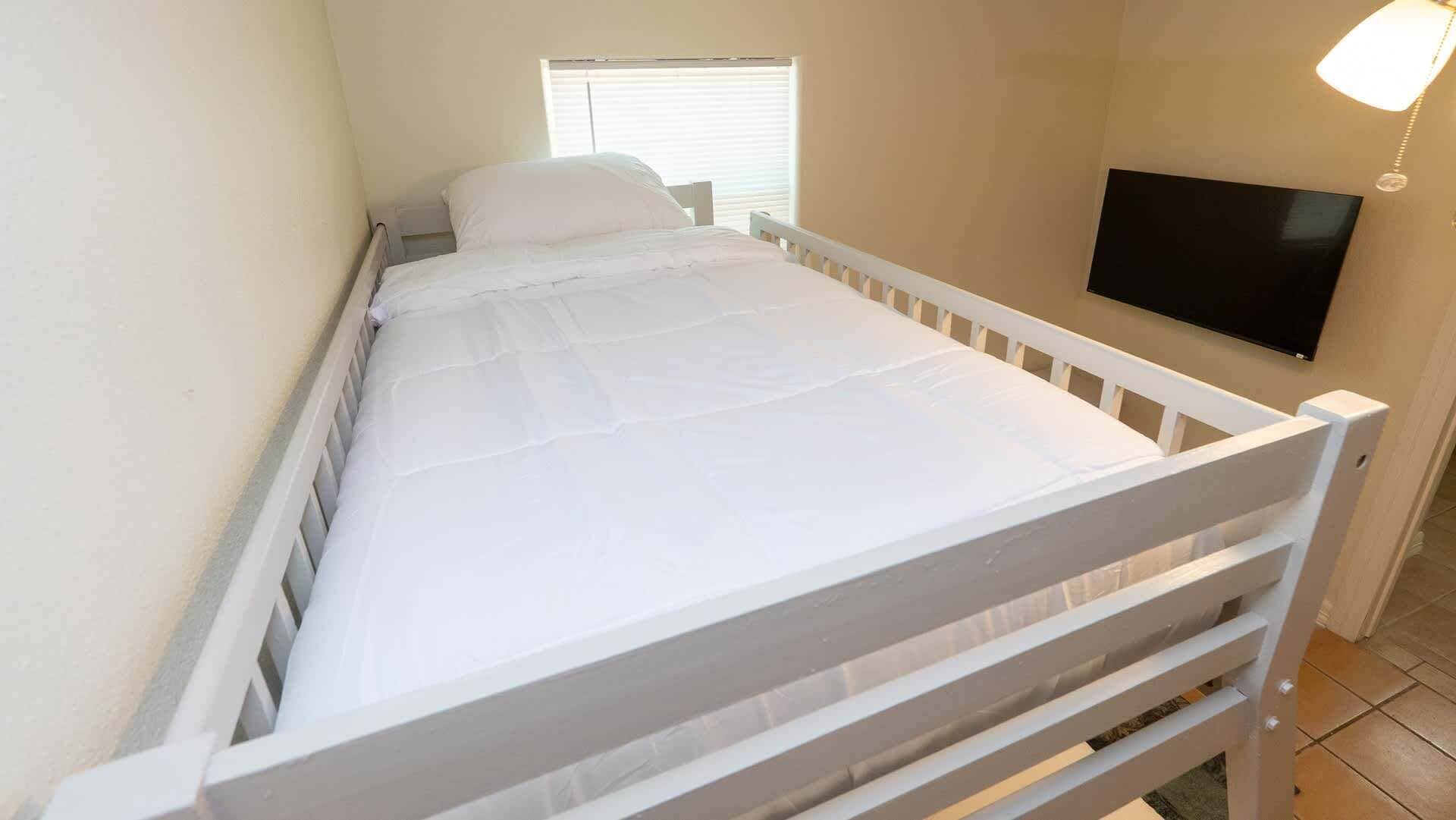 Uma cama de solteiro com colchão e cabeceira brancos, uma janela com persianas e uma televisão de tela plana fixada na parede.