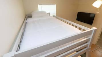 Uma cama de solteiro com colchão e cabeceira brancos, uma janela com persianas e uma televisão de tela plana fixada na parede.