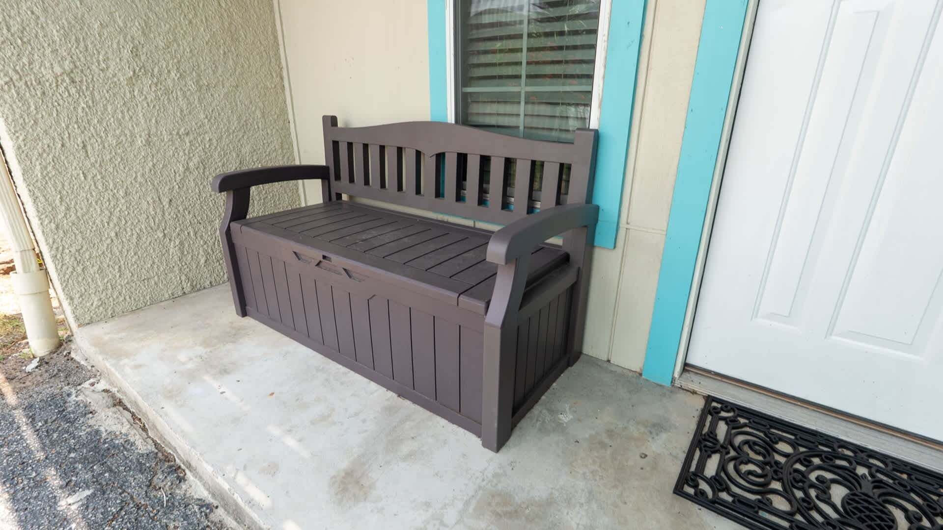 A bench with a slatted seat and backrest, positioned on a concrete porch with a textured wall and a white door with a blue trim.