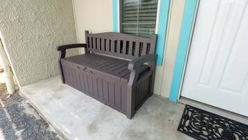 A bench with a slatted seat and backrest, positioned on a concrete porch with a textured wall and a white door with a blue trim.