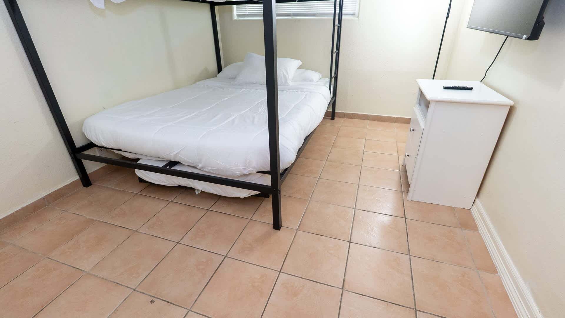 A single bed with a metal frame and white bedding in a room with tiled flooring and a small white cabinet.