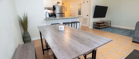 Un comedor con mesa de madera y bancos, cocina al fondo y un televisor en la pared.