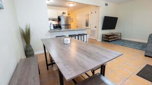 Un comedor con mesa de madera y bancos, cocina al fondo y un televisor en la pared.