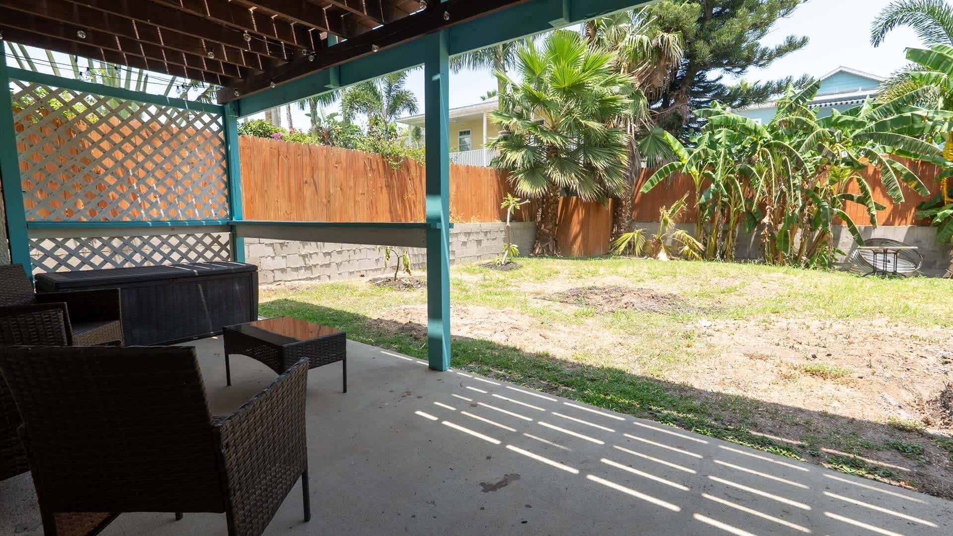 Une terrasse extérieure avec des meubles en osier, une clôture en bois et une vue sur une cour arrière agrémentée de palmiers et d’une maison.