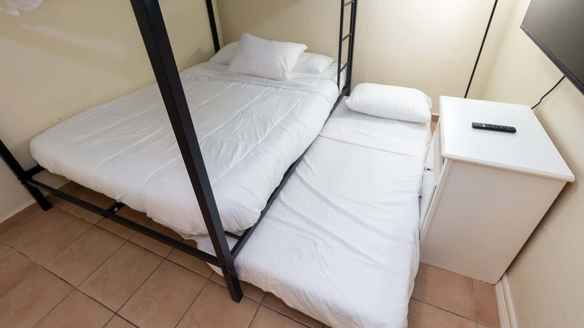 Un lit simple avec un cadre noir, un matelas blanc et un oreiller blanc. Il y a une petite table de chevet blanche sur laquelle se trouve une télécommande.