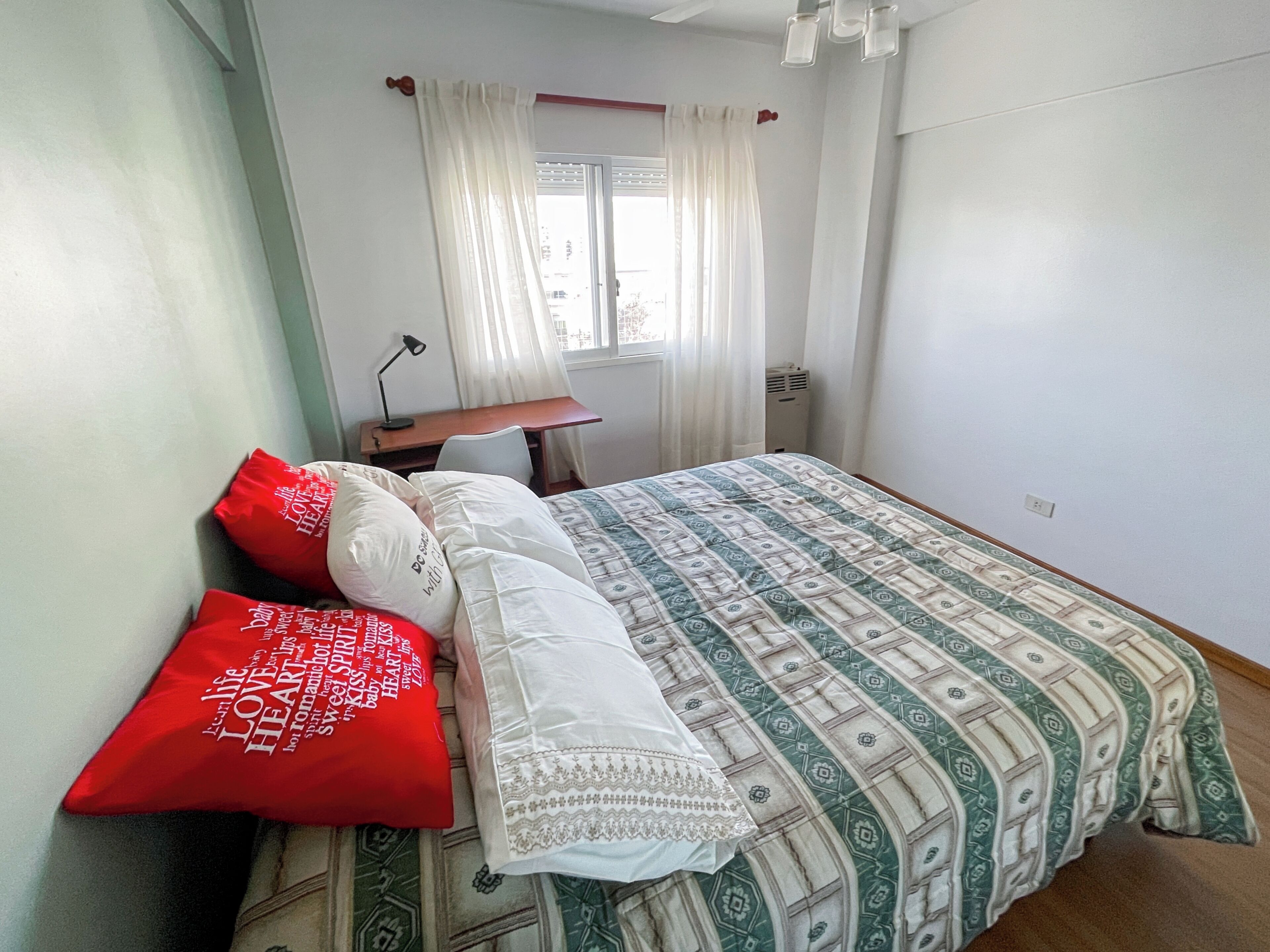 Quarto com cama, escrivaninha, cadeira, janela com cortinas e ventilador de teto.
