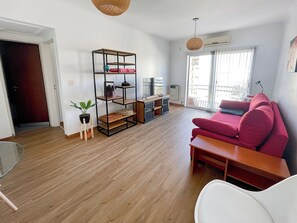 Uma sala de estar moderna com um sofá vermelho, piso de madeira, uma estante de metal preta e uma grande janela com vista para edifícios.