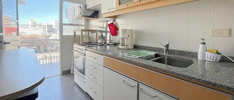 Une cuisine moderne avec des meubles blancs, un plan de travail en granit, une cuisinière à gaz, une machine à café et une vue sur la ville grâce à de grandes fenêtres.