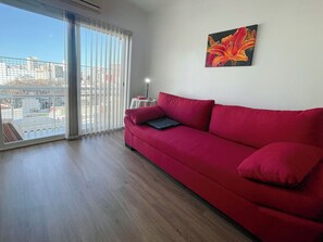 Um sofá vermelho numa sala com uma janela grande, uma mesa pequena e um quadro na parede.