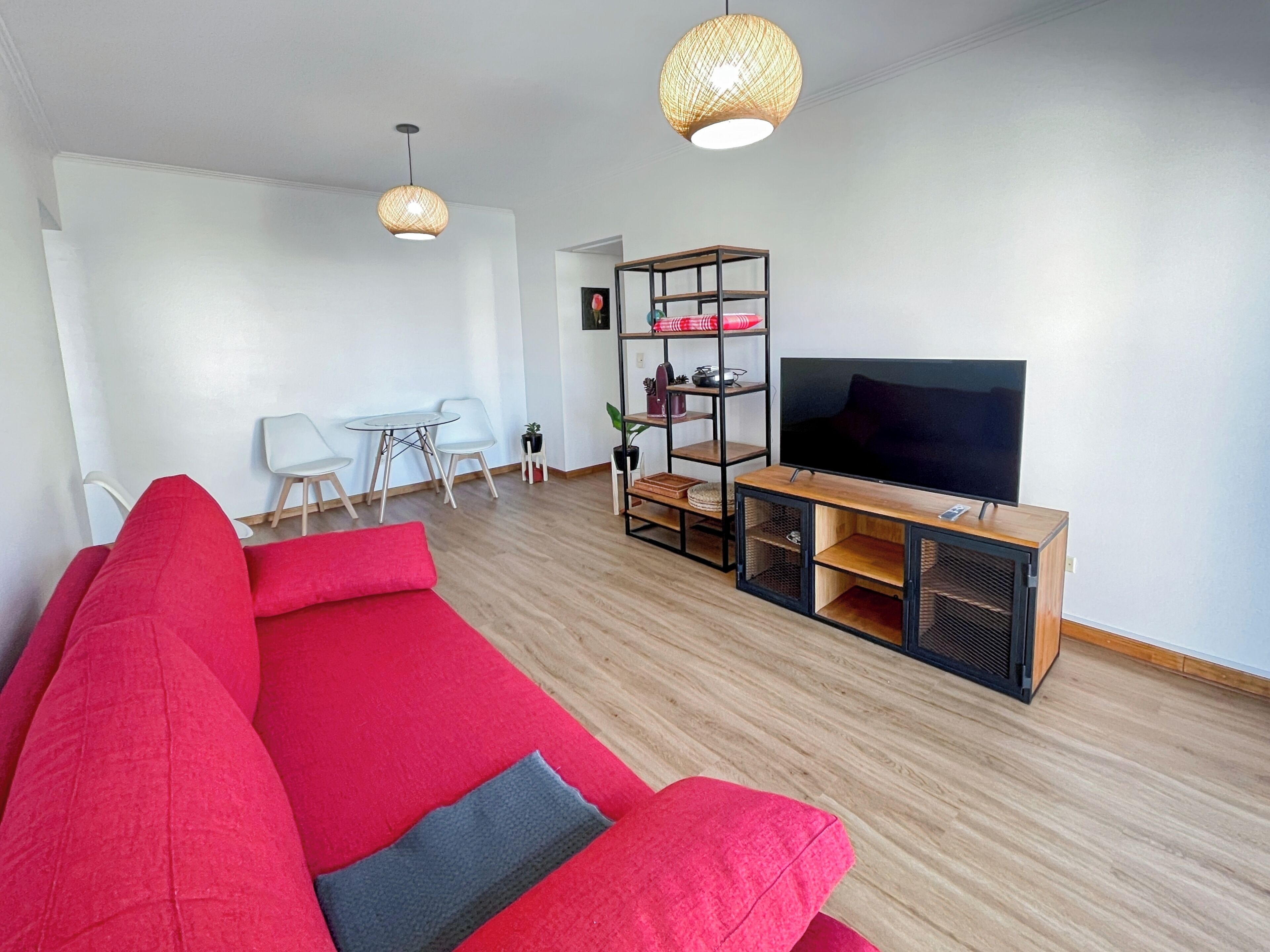 Uma sala de estar moderna com um sofá vermelho, uma TV sobre um suporte de madeira, e uma área de jantar branca com cadeiras.