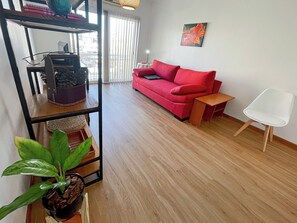 Uma sala de estar com um sofá vermelho, uma cadeira branca, uma mesa de centro de madeira, uma estante de metal preta com plantas e um quadro na parede.