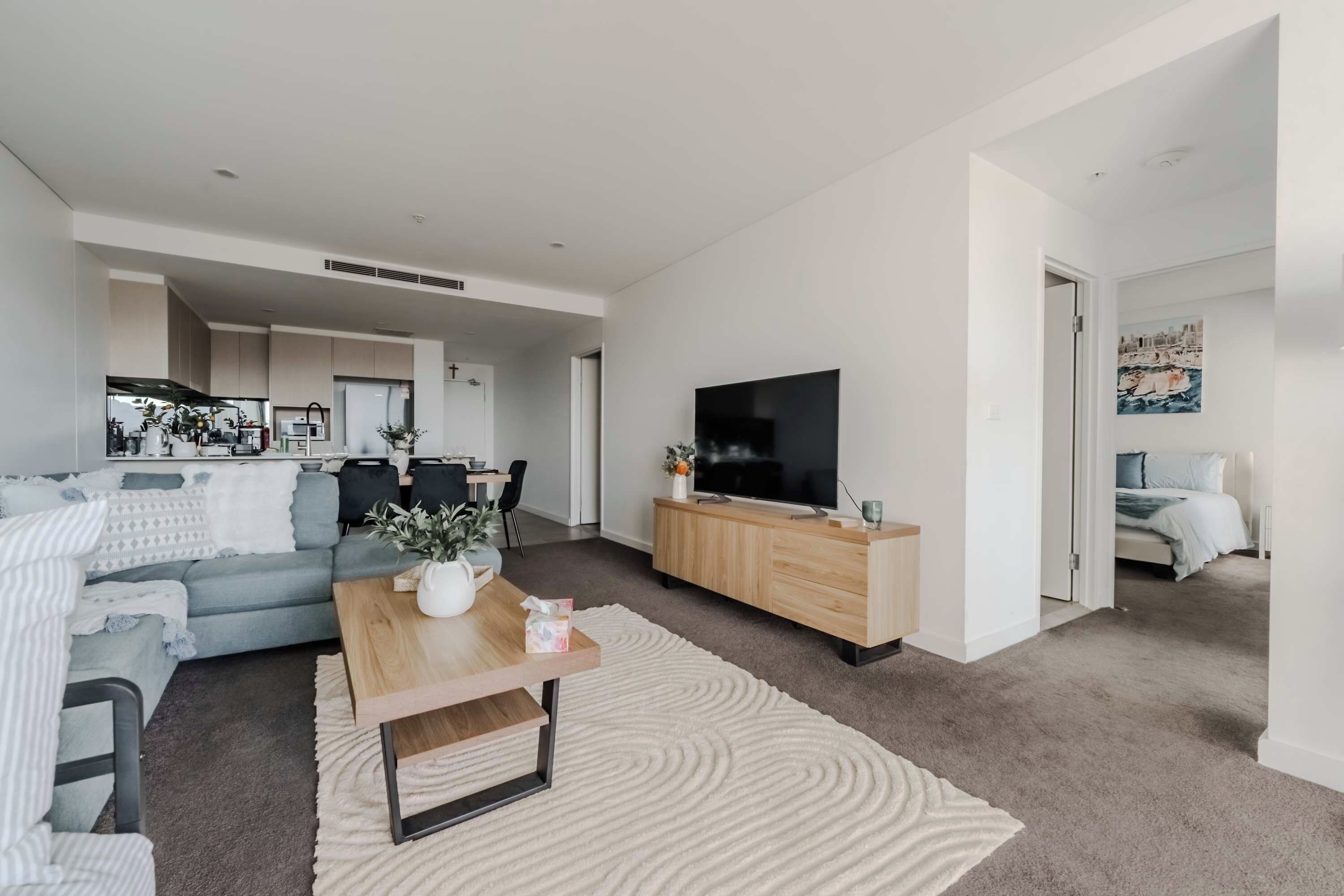 Ein modernes Wohnzimmer mit einer Couch, einem Couchtisch und einem Fernseher. Im Hintergrund befindet sich eine Küche mit Schränken und Geräten.