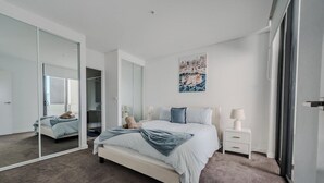 Une chambre à coucher moderne avec un grand lit, une table de chevet avec une lampe, un miroir et un tableau représentant un paysage urbain accroché au mur.