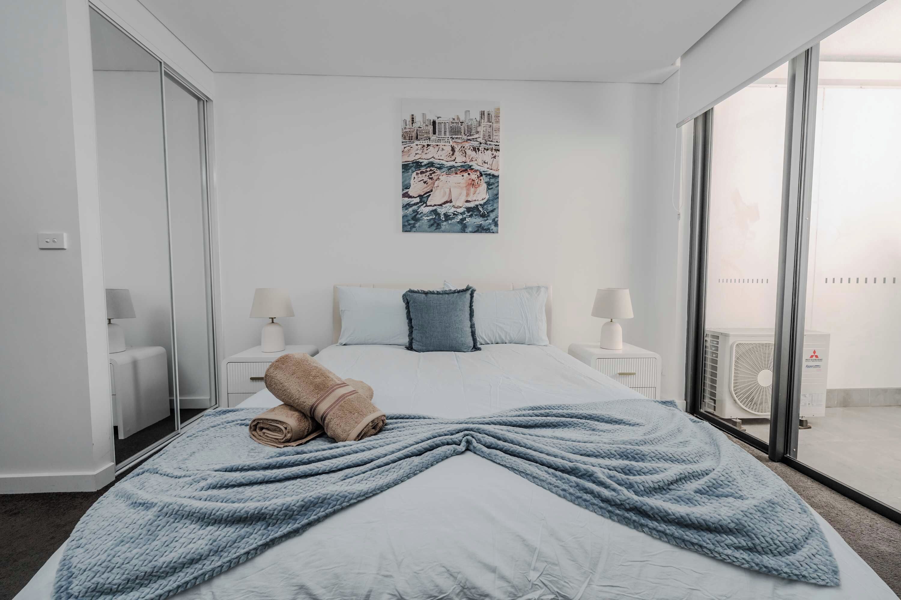 Une chambre d’hôtel avec un lit, deux tables de chevet, une grande fenêtre et un tableau représentant un paysage urbain accroché au mur.