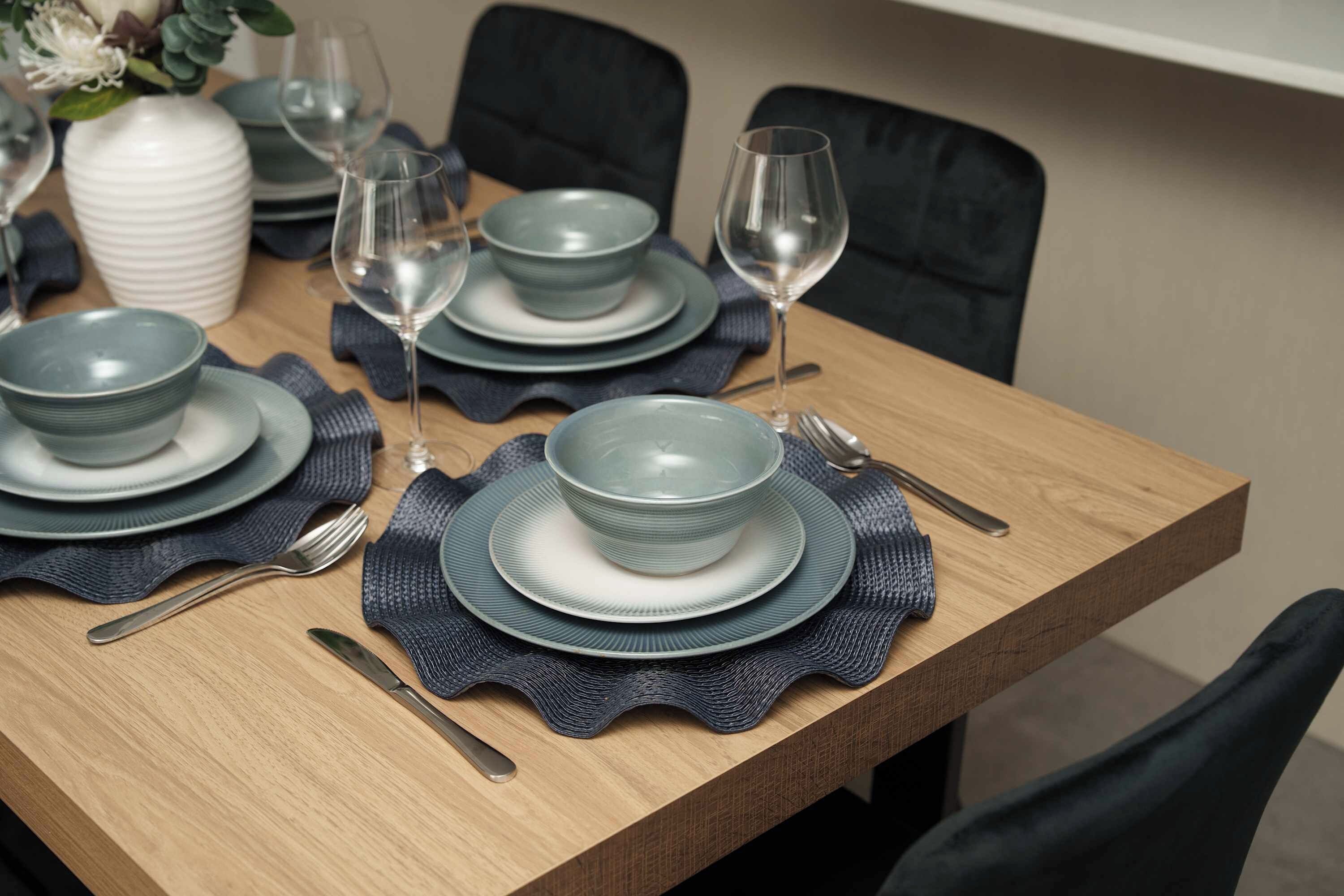 Een eettafel gedekt met borden, kommen, bestek en wijnglazen, met een vaas en een potplant op de achtergrond.