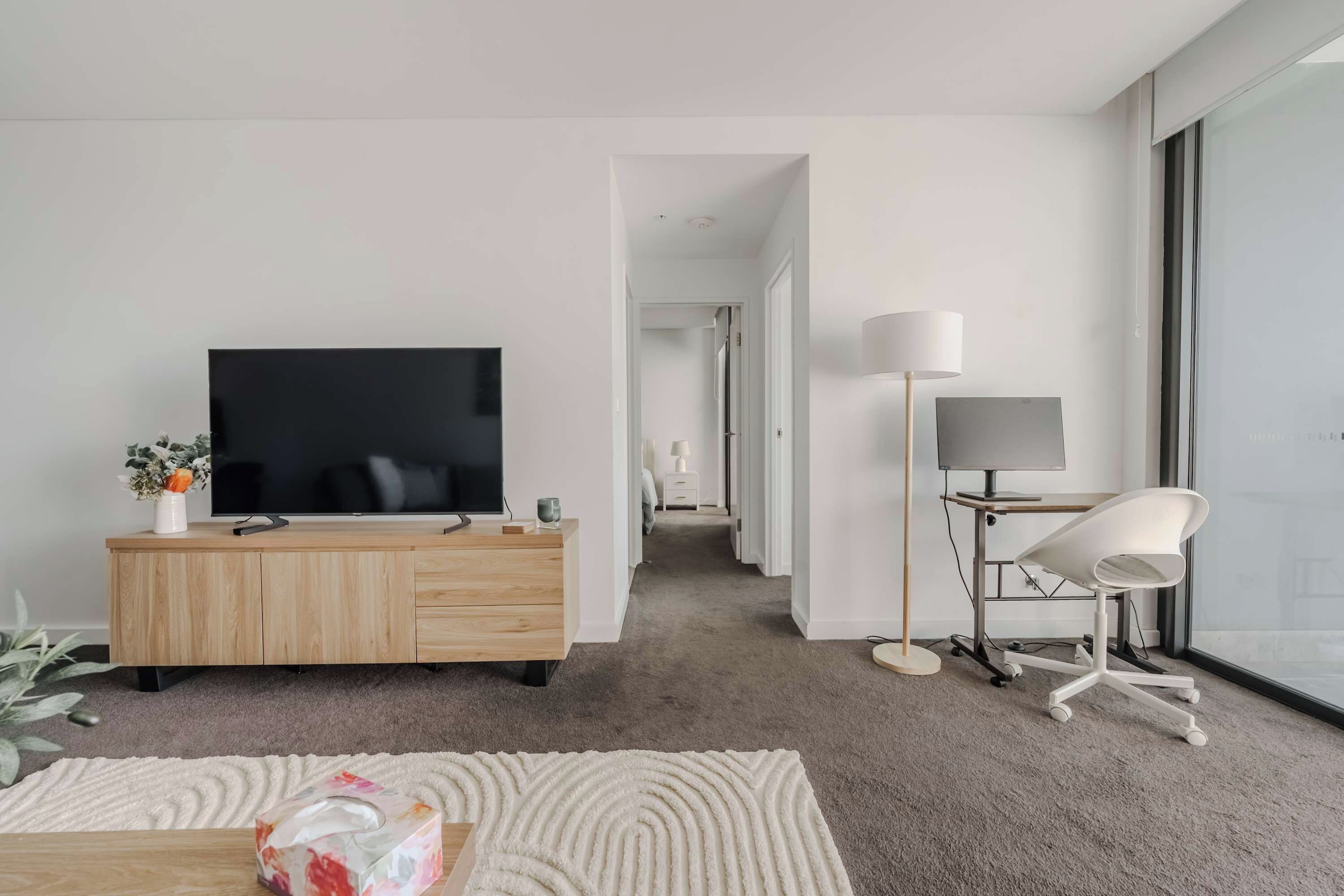 Un salon moderne avec un téléviseur à écran plat, un meuble en bois, un bureau avec une lampe et une chaise blanche.