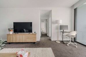 A modern living room with a flat-screen TV, a wooden cabinet, a desk with a lamp, and a white chair. - Trinitus | Mountain View | Parking | Central (Wollongong)