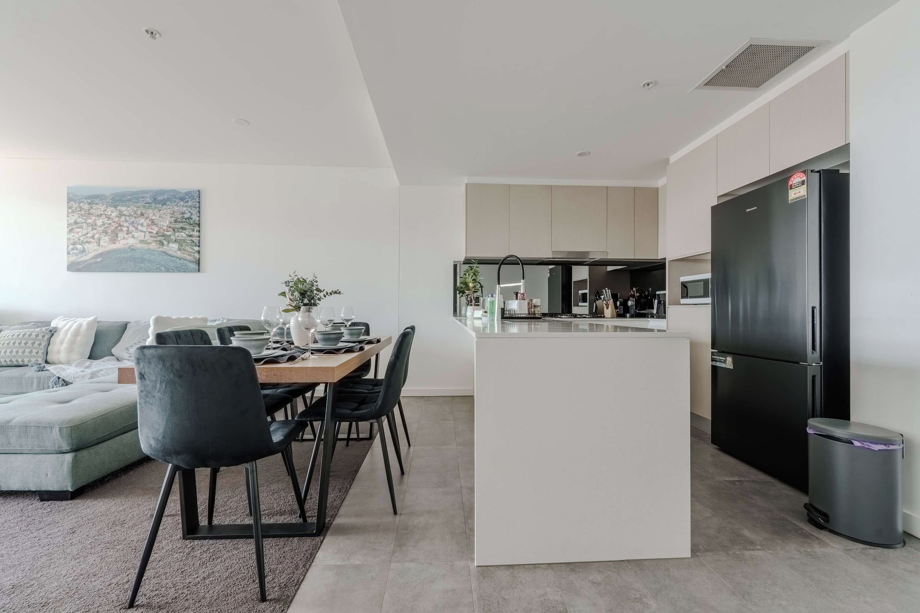 Een moderne keuken met een zwarte koelkast, een zithoek met een tafel en stoelen, en een woonkamer met een bank en een schilderij aan de muur.