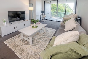A modern living room with a flat-screen TV, a white coffee table, a grey sofa with cushions, and a patterned rug. - Spiritus | Mountain View | Parking | Central (Wollongong)