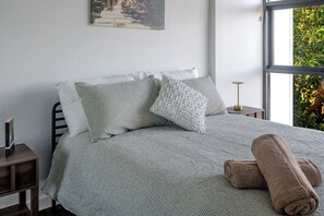 Ein ordentlich bezogenes Bett mit einer grauen, gemusterten Tagesdecke, einem weißen Kopfkissen und einem aufgerollten braunen Handtuch daneben.