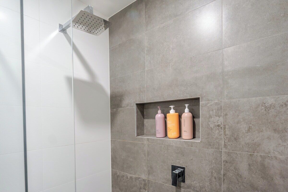 A bathroom with a shower, a shelf with three bottles, and a black faucet.