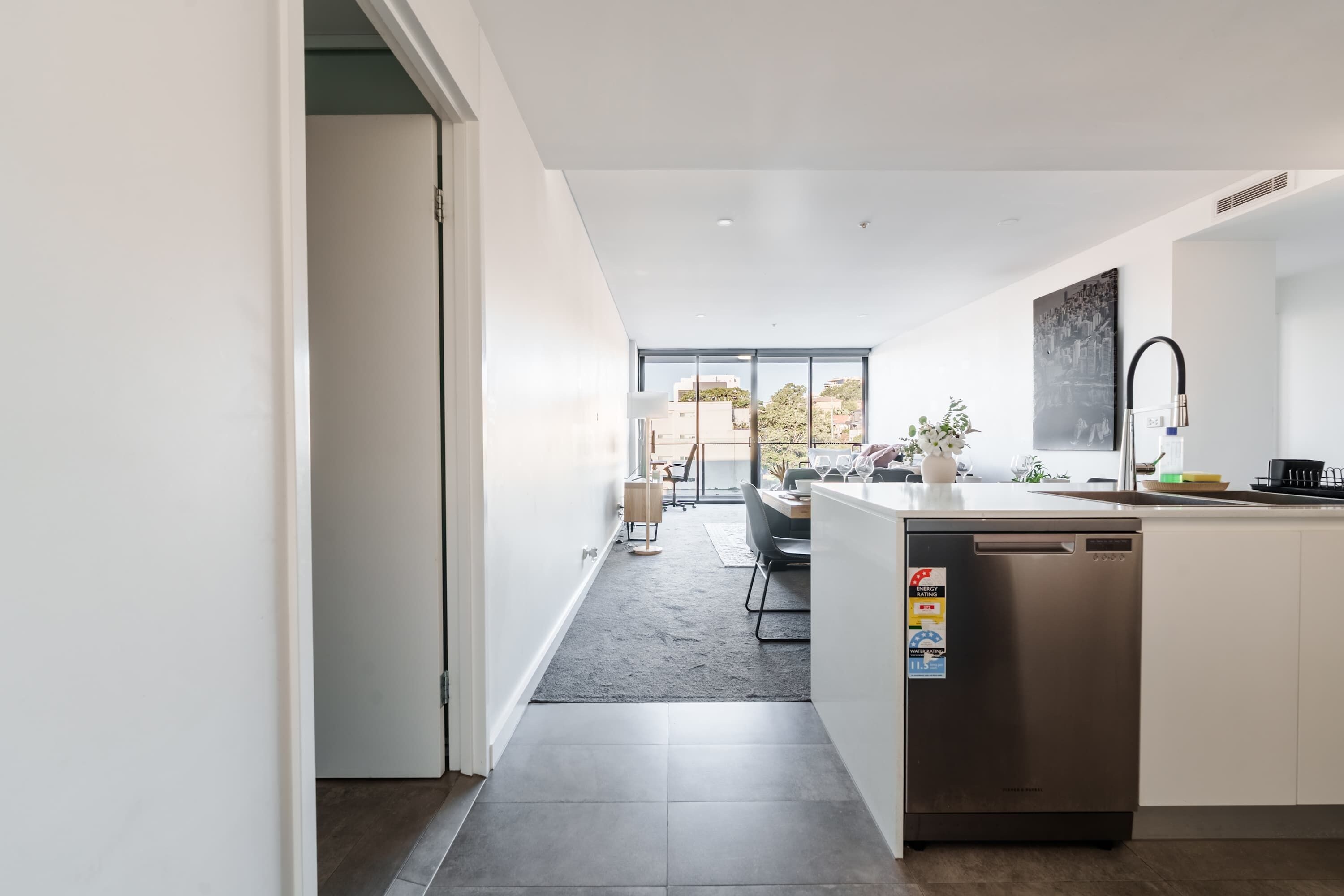 A modern kitchen with a dishwasher, a dining area with a table and chairs, and a view of the outdoors through large windows.