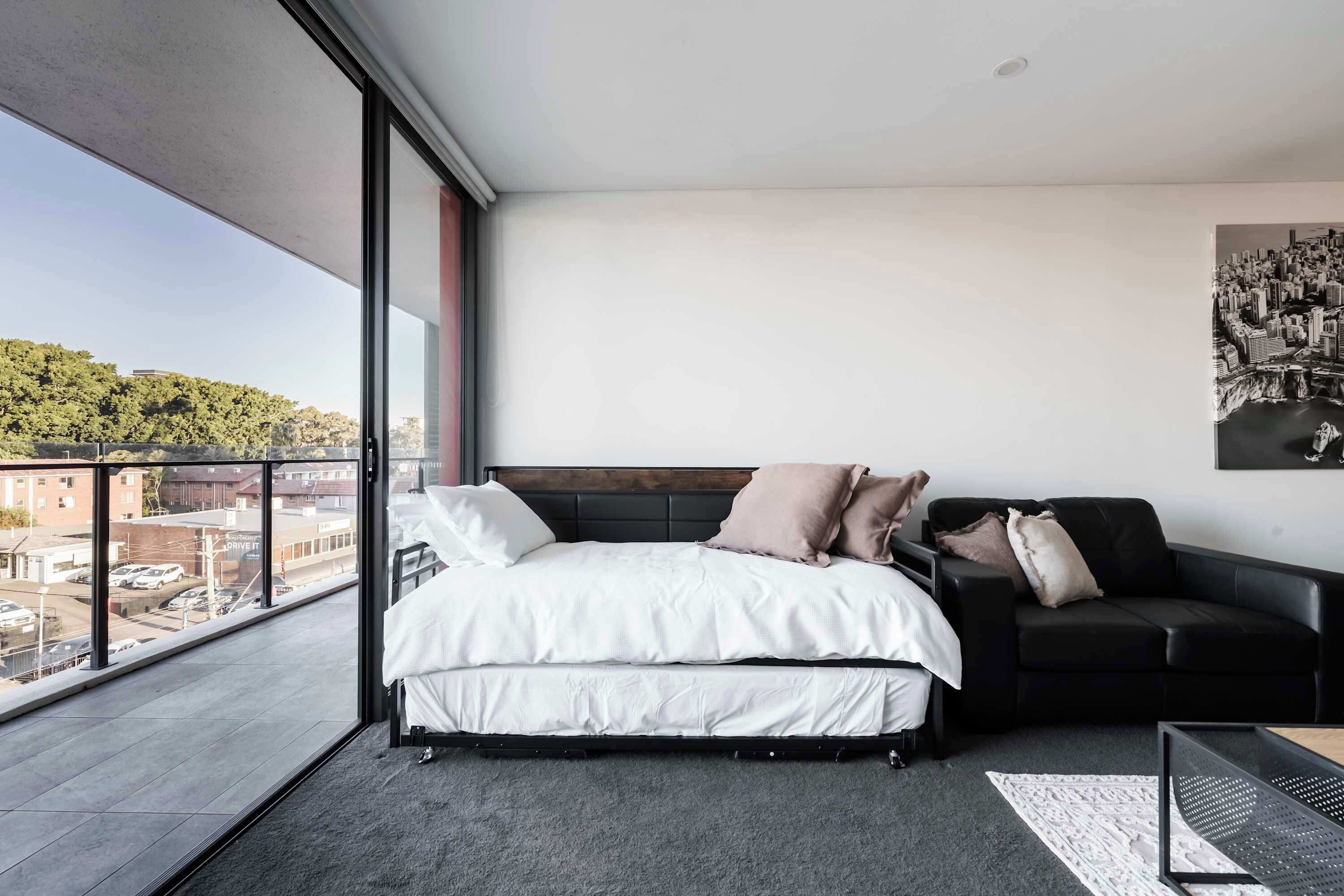 A modern bedroom with a large bed, a black sofa, and a cityscape view through large windows.