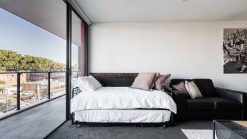 A modern bedroom with a large bed, a black sofa, and a cityscape view through large windows.