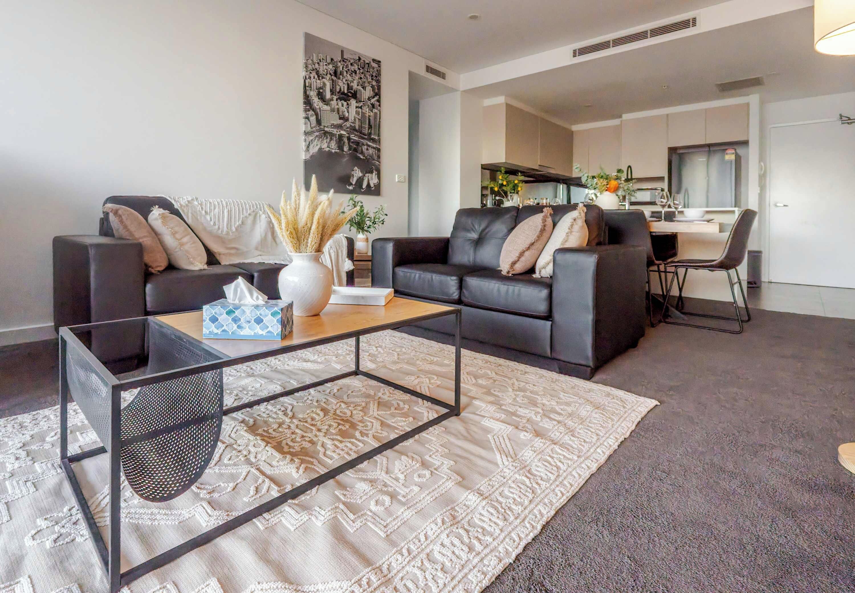 A modern living room with a glass coffee table, two sofas, and a dining area in the background.