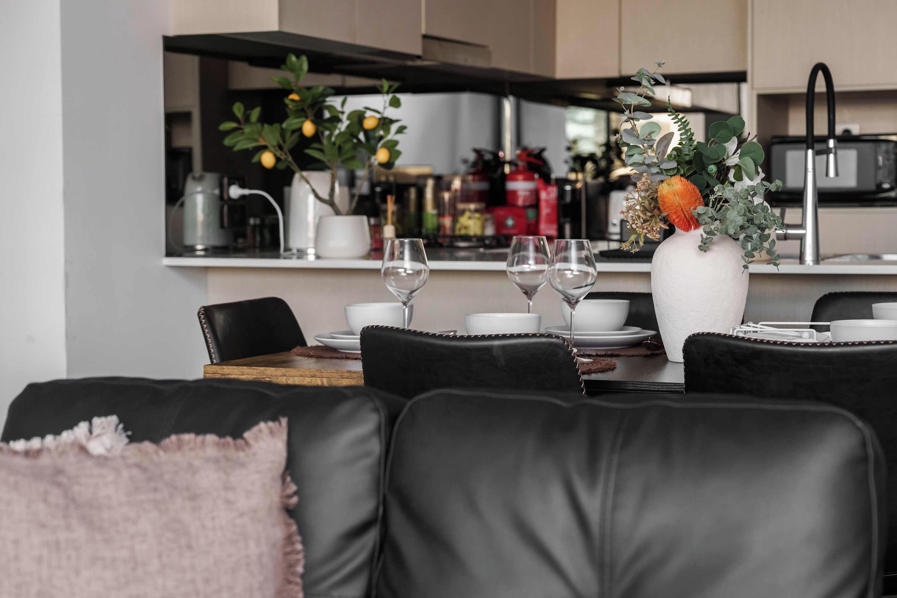 A modern dining area with a black leather couch, a white vase with flowers, and a kitchen in the background.
