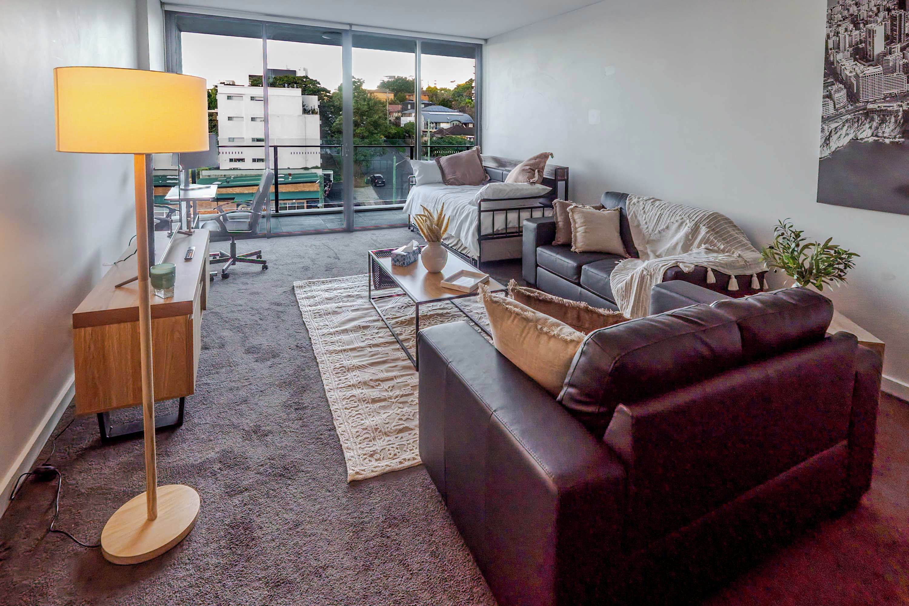 Een moderne woonkamer met een bank, een bureau, een lamp en een stadszicht door grote ramen.