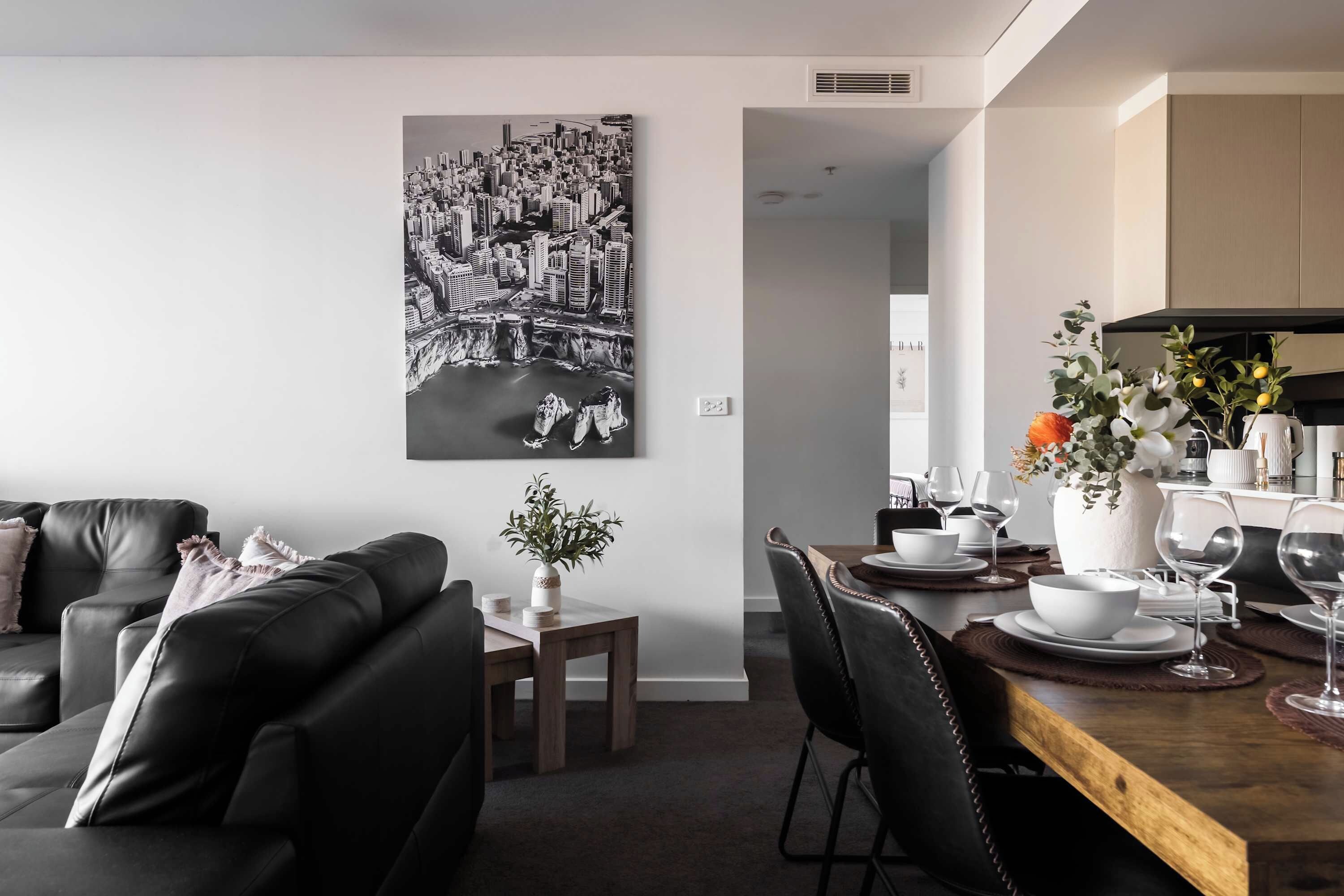 Een moderne woonkamer met een zwarte leren bank, een eethoek met een houten tafel gedekt voor twee personen, en een stadsgezichtschilderij aan de muur.