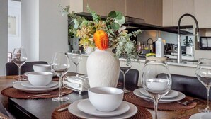 A dining table set with white plates, bowls, and wine glasses, a vase with flowers, and a kitchen in the background. - Filioque | Mountain View | Parking | Central (Wollongong)