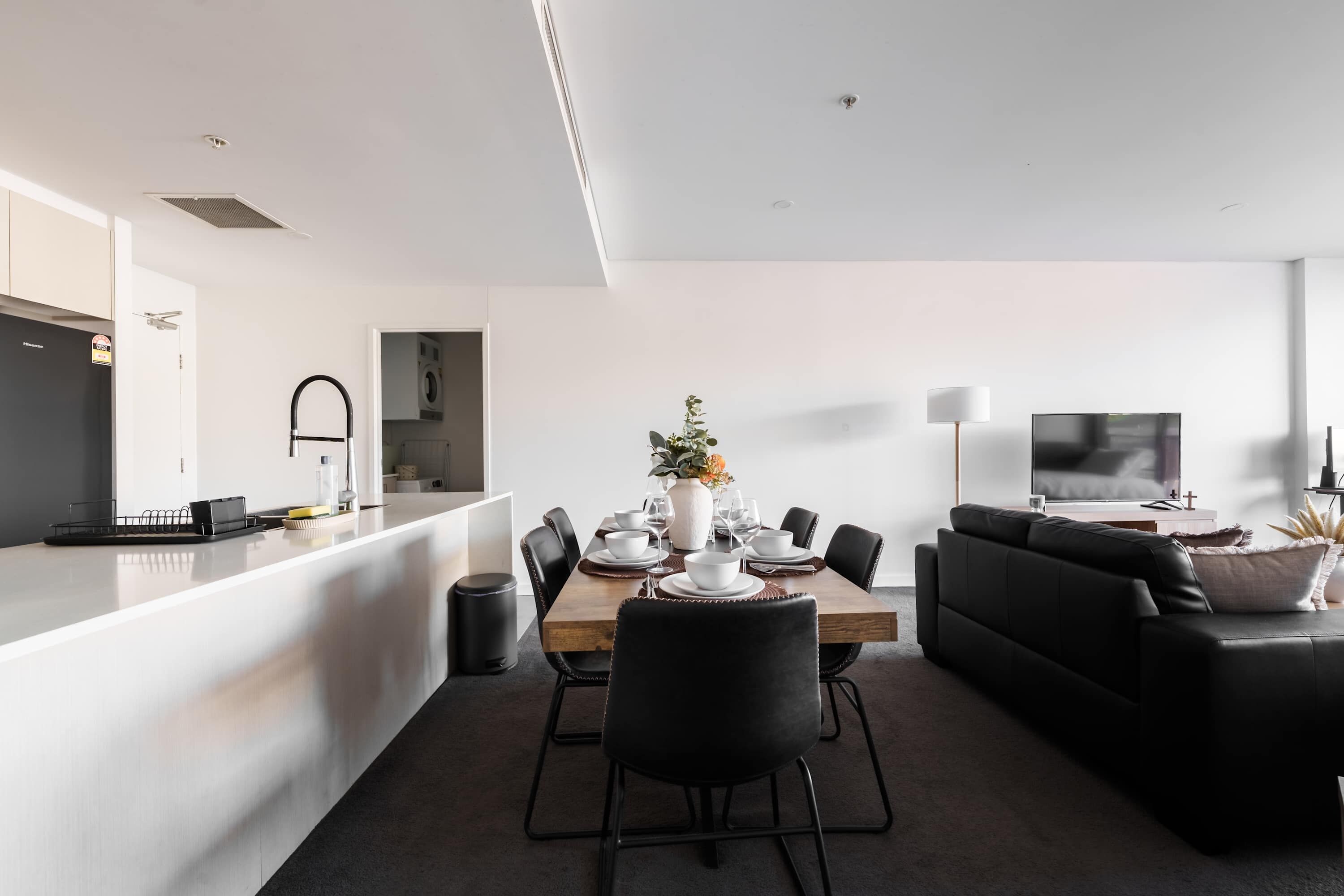 A modern kitchen with a built-in oven, a dining area with a wooden table and black chairs, and a living area with a black sofa and a TV.