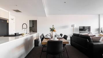 A modern kitchen with a built-in oven, a dining area with a wooden table and black chairs, and a living area with a black sofa and a TV.