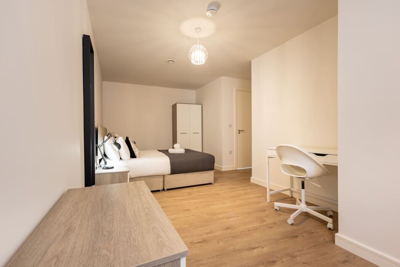 A bedroom with a bed, bedside table, lamp, wardrobe, and a desk with a chair.
