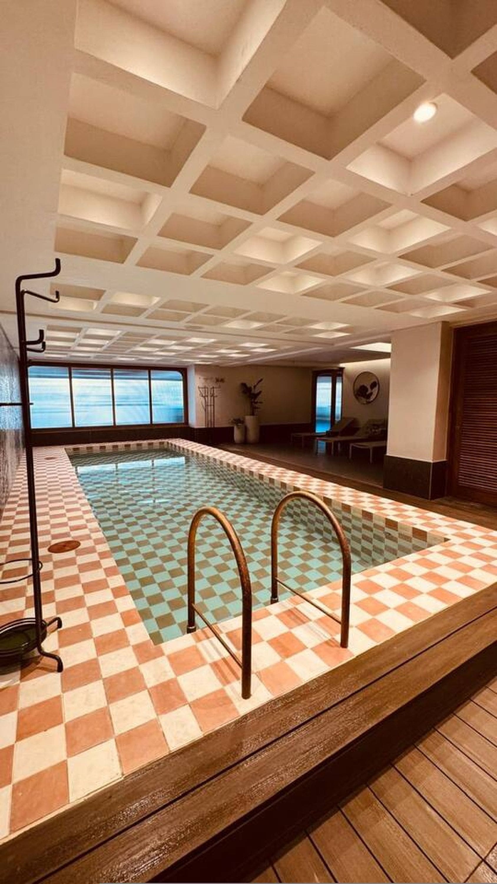 Una piscina interior con baldosas de diseño ajedrezado, una plataforma de madera e iluminación empotrada en el techo.