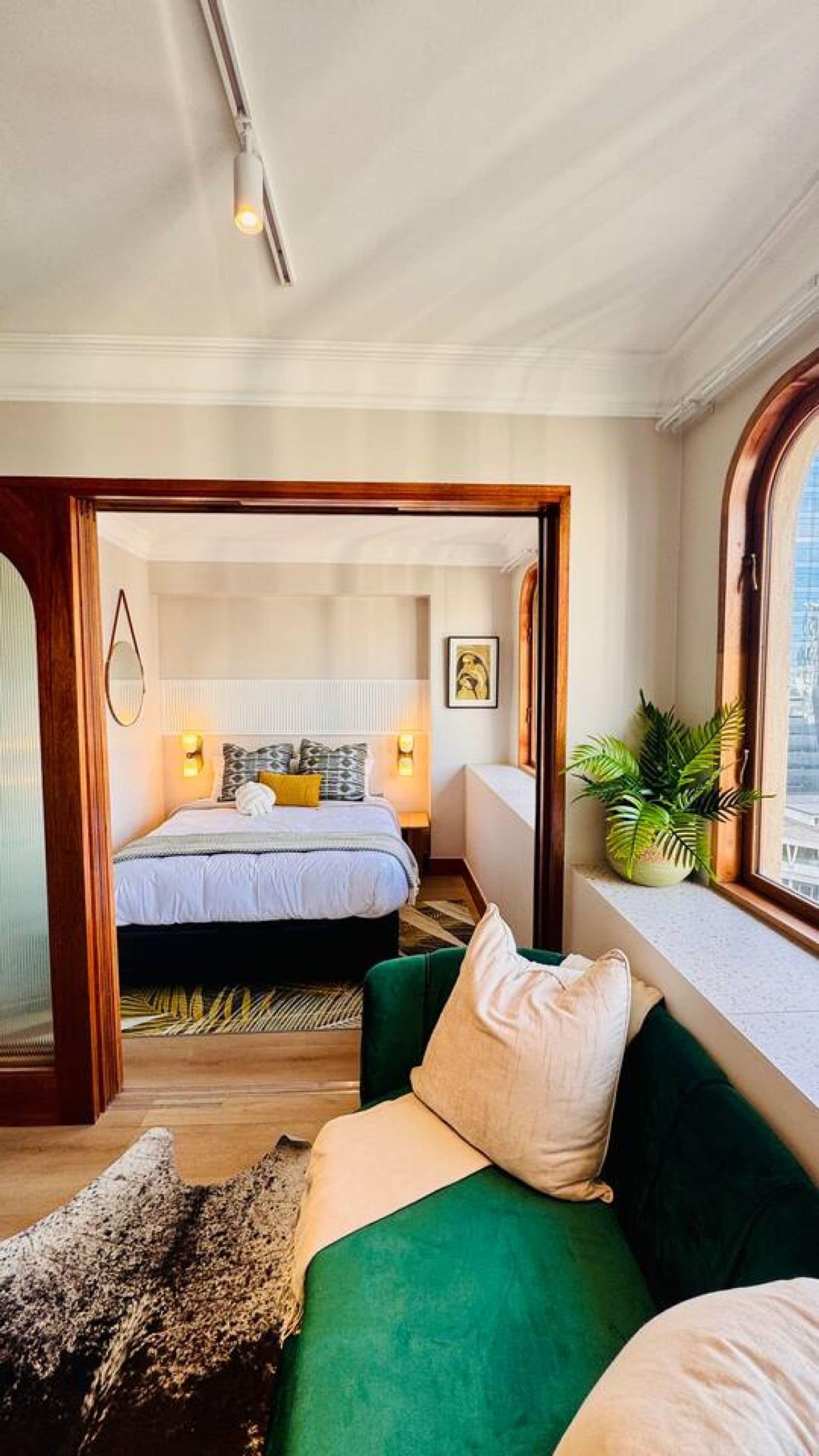 Quarto com cama, sofá verde, janela com planta e porta que dá para outro cômodo.