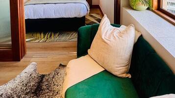 A bedroom with a bed, a green sofa, a window with a plant, and a door leading to another room.