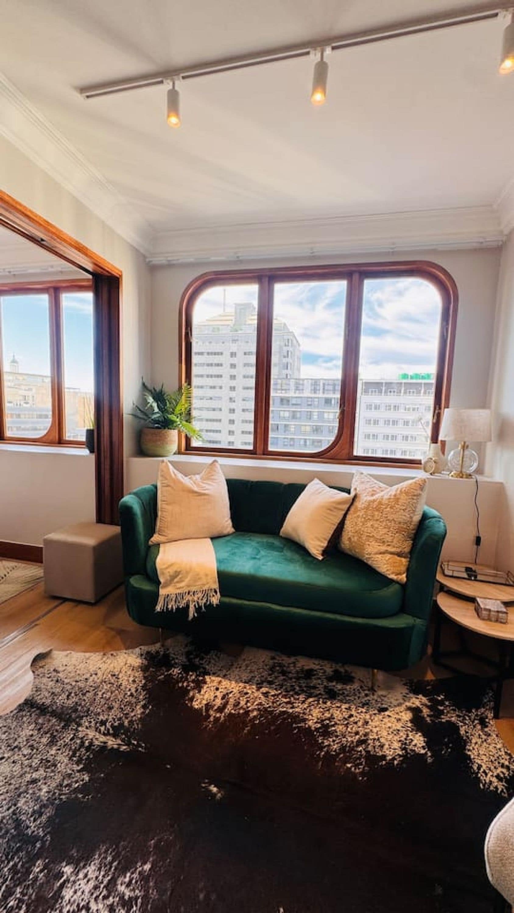 A room with a green sofa, a black and white rug, a potted plant, and a view of buildings through large windows.
